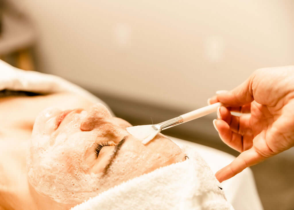 A facial treatment in progress with a client relaxing while a professional applies a mask with a brush.
