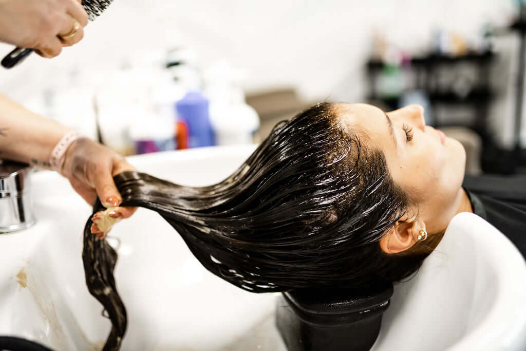Woman receiving a hair treatment at a salon with shiny, wet hair while relaxing in a sink.